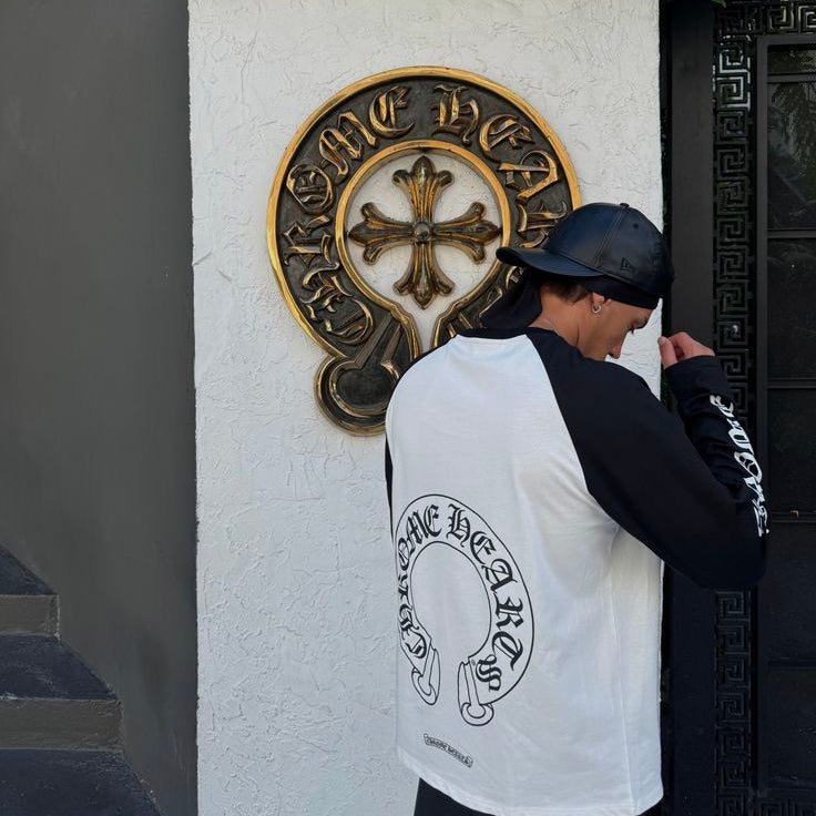 A person wearing a black and white raglan long-sleeve shirt with a large Chrome Hearts logo printed on the back, standing in front of a white wall with a gold-toned Chrome Hearts emblem.