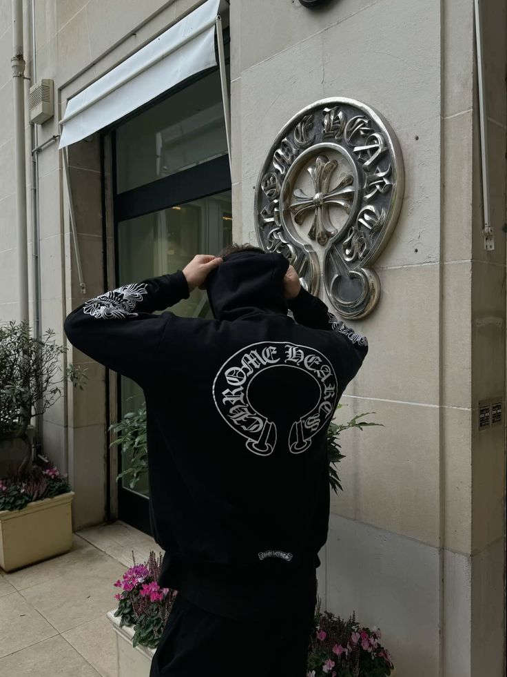 Person wearing a black hoodie with a logo, standing in front of a decorative wall piece on a building.