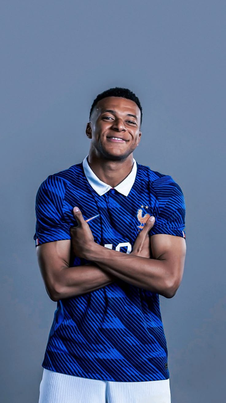A man wearing a blue patterned soccer jersey with white shorts, posing with his arms crossed against a solid gray background.