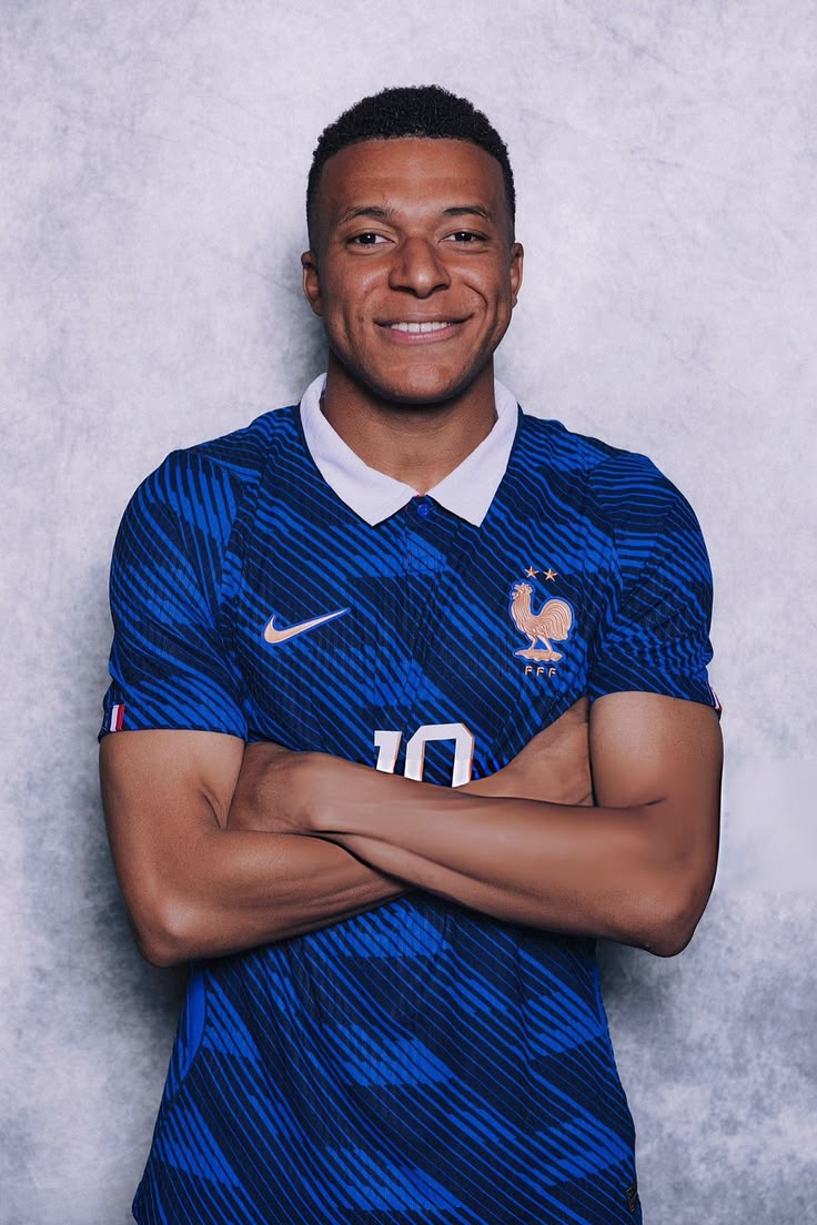 A smiling man wearing a blue France football jersey with a white collar and Nike logo, posed with arms crossed against a textured light gray background.