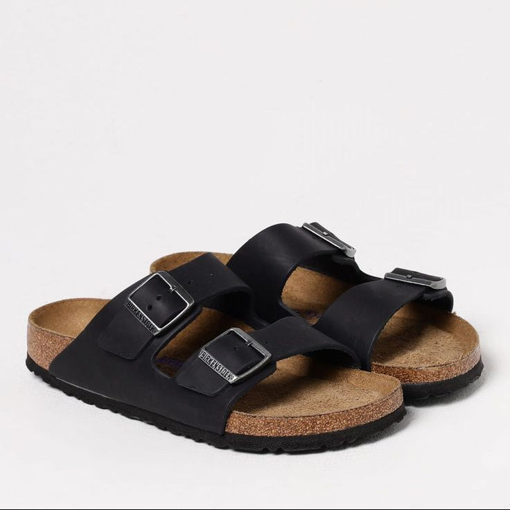A pair of black double-strap slide sandals with adjustable metal buckles and a natural cork footbed, shown on a clean white background.