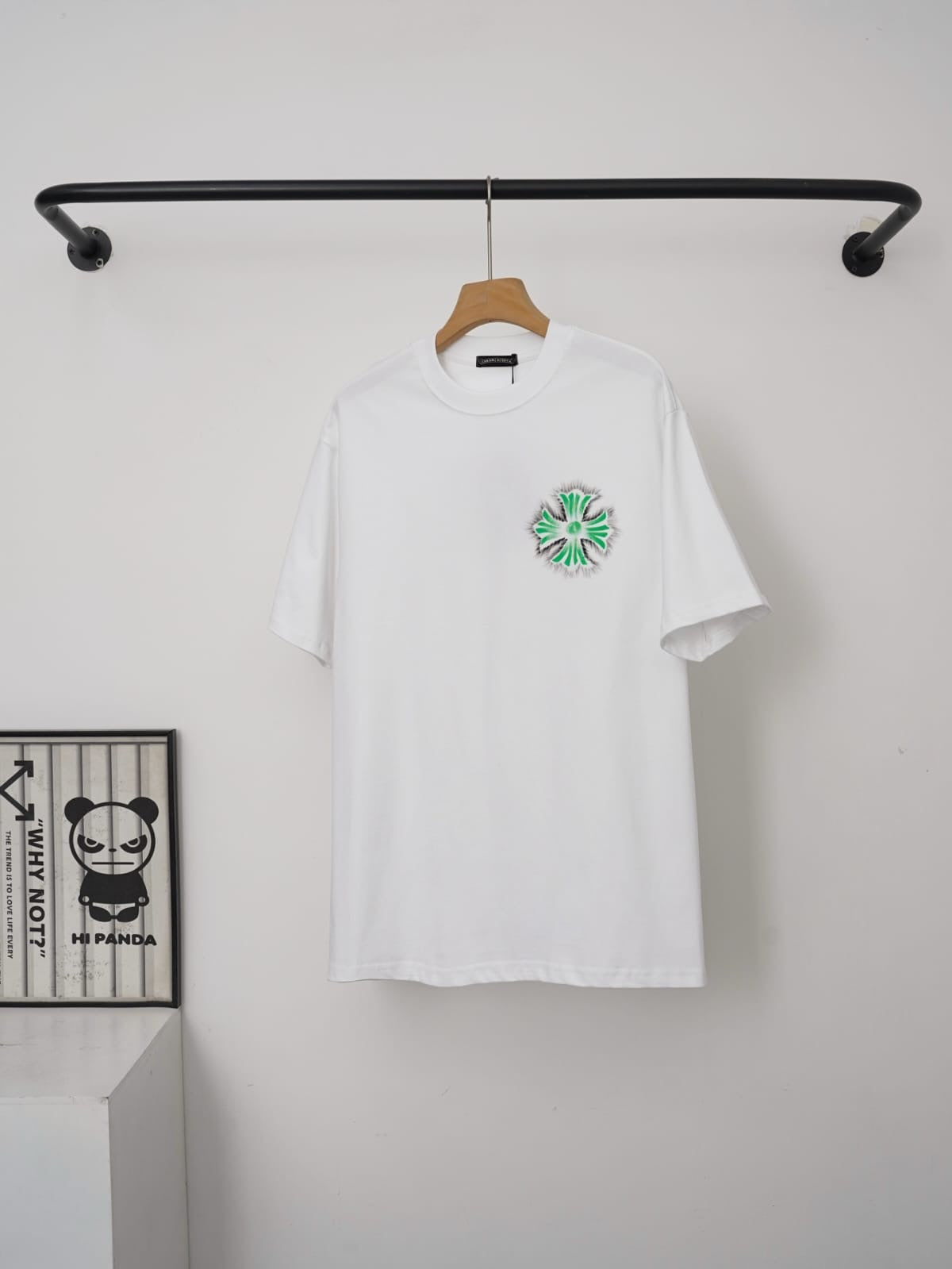 White t-shirt with a green and red logo on a hanger against a white wall.