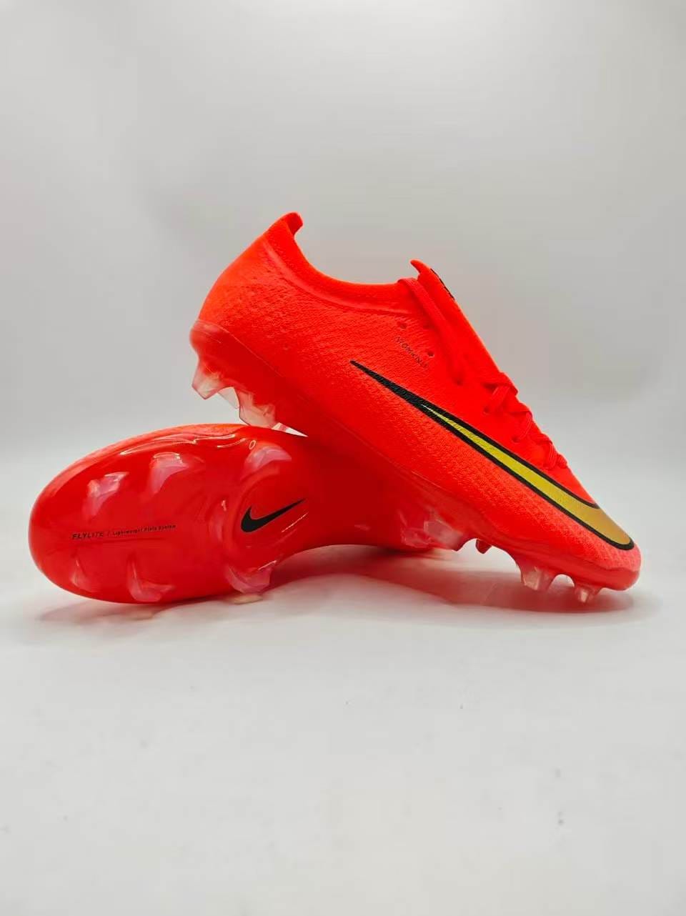 A pair of Nike soccer cleats in a vibrant neon orange-red colorway, featuring a black Swoosh with a yellow outline accent, shown against a solid black background.